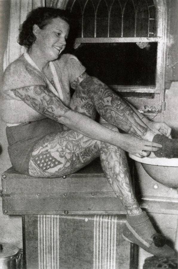 A young white woman with fully tattooed legs and arms tattooed to her elbows sits while she puts on a sock.