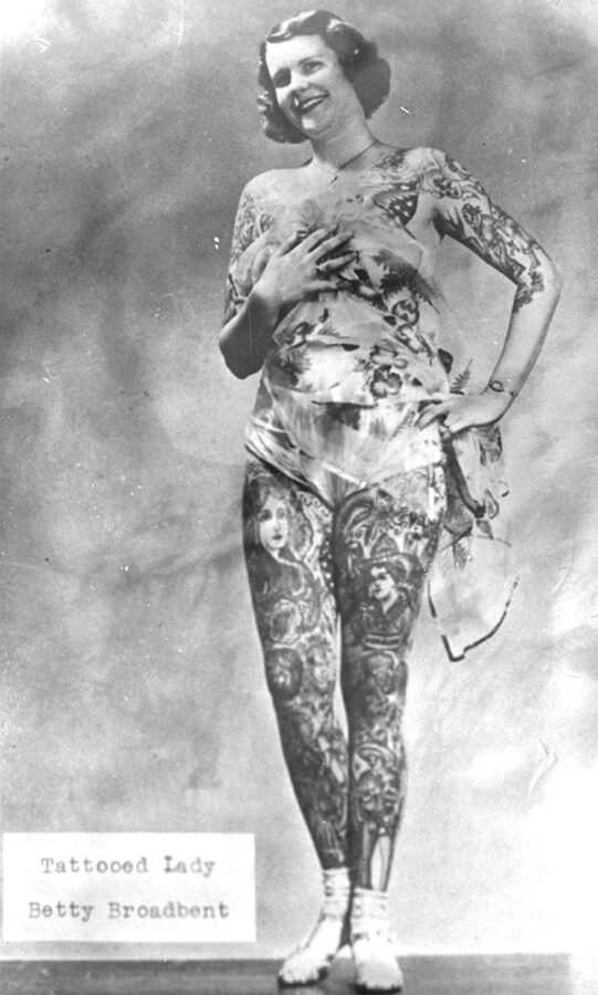 A young, heavily tattooed white woman stands holding a cloth covering over her chest. In the bottom corner are the words "Tattooed Lady Betty Broadbent."
