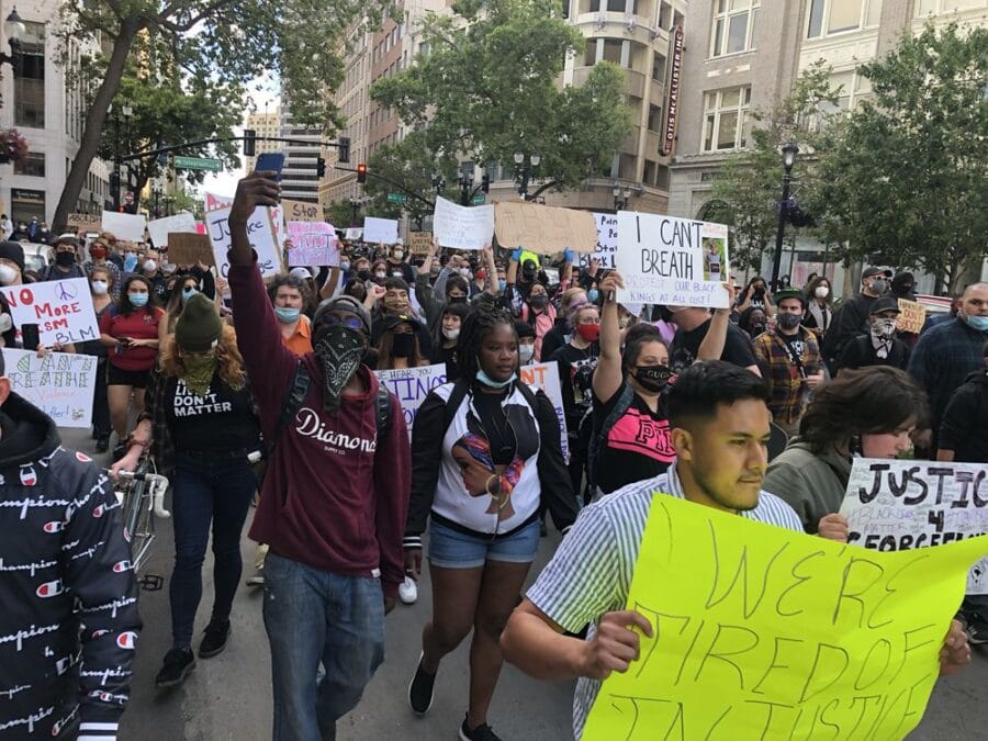 A large group of people march in a protest. Some are holding signs, which read "We're tired of injustice" and "I can't breathe," among other slogans.