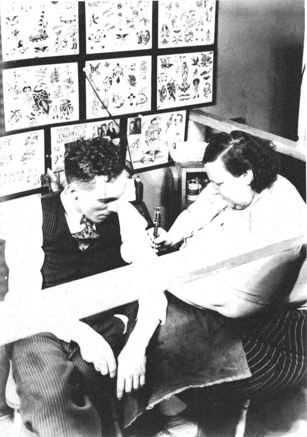 A fat white woman tattoos a thin white man's arm. On a nearby wall are sheets of paper with different tattoo designs.