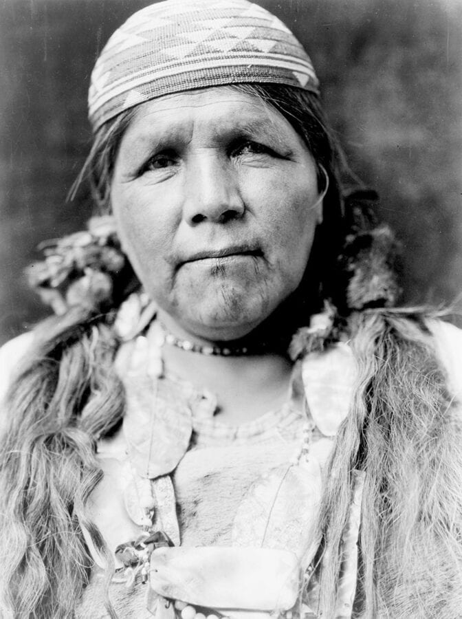 An older Hupa woman wears a basket cap and has faint tattoo markings on her chin.