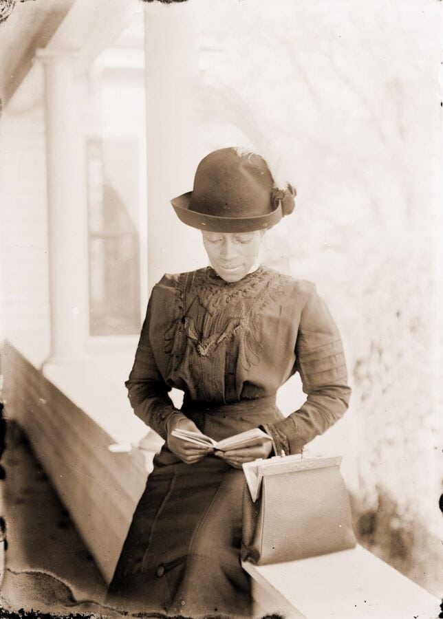 A young African American woman wears a nice dress and hat and sits on a porch. She is reading a book and has her handbag next to her.