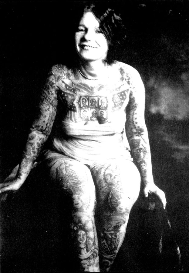 A fully-tattooed young white woman with a bob haircut smiles at the camera as she leans forward in a seated position. She is wearing a bodysuit, and her many tattoos are clearly visible.
