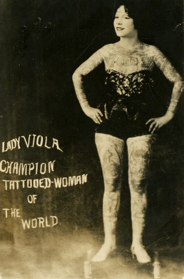 A heavily-tattooed young white woman wears a sparkly bodysuit and heels. She stands with her hands on her hips and smiles for the camera. The text on the photo reads, "Lady Viola Champion Tattooed-Woman of the World."