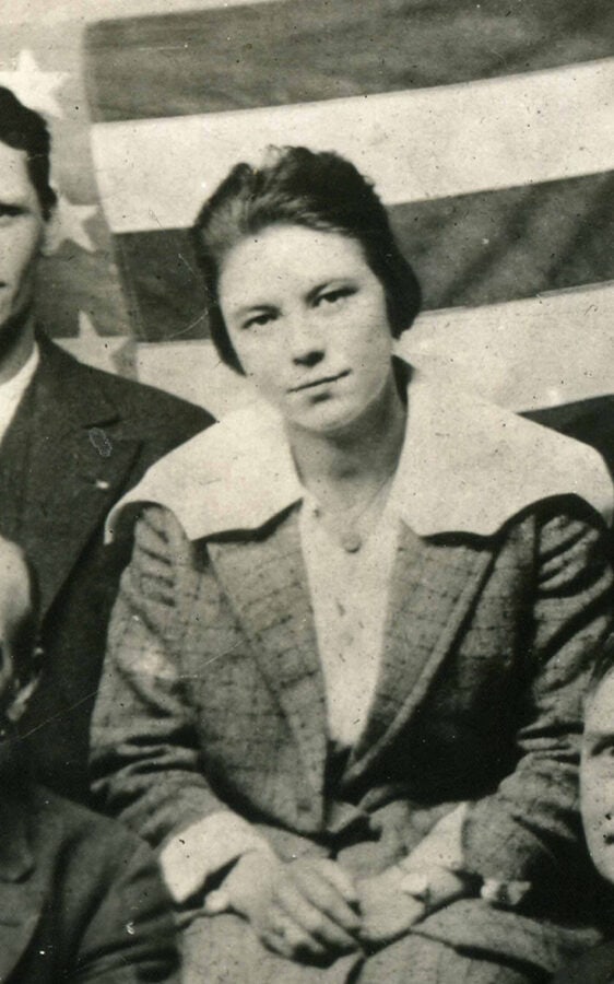 A young white woman sits with her hands clasped in her lap in a group photo, although the photo crops out the other faces. An American flag is used as the photo's backdrop.