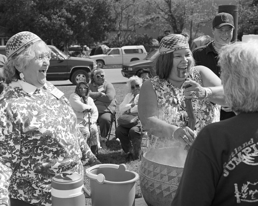 At a gathering, a Native woman wearing a basket cap and a chin tattoo makes acorn soup. Another, older Native woman with a basket cap looks on. Both are laughing and wear modern dress.