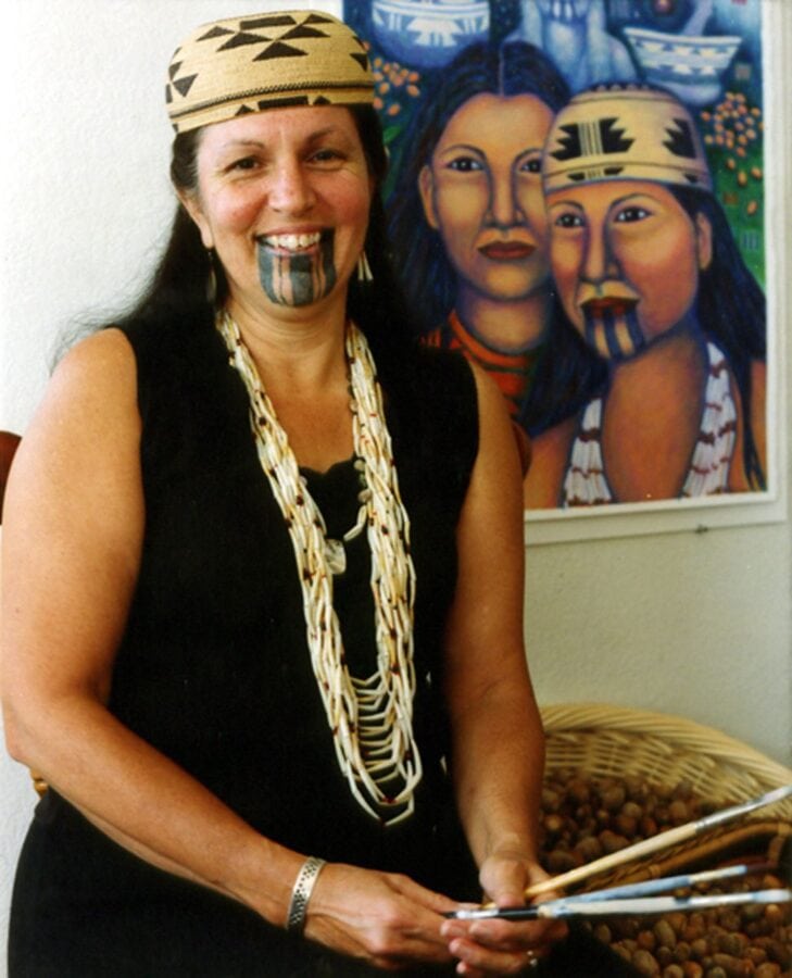 A Native woman wears a shell bead necklace and basket hat. She has three thick black lines as a chin tattoo. A painting showing two Native women, one with a chin tattoo, is behind her.