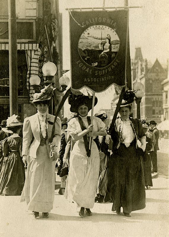 Three women hold a flag that says "California Equal Suffrage Association" while marching outside.