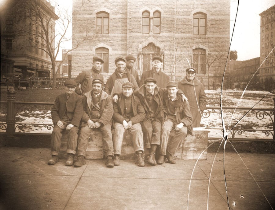 A photo shows one black man and nine white men in front of an old brick building. There is snow on the ground and no leaves on the trees.