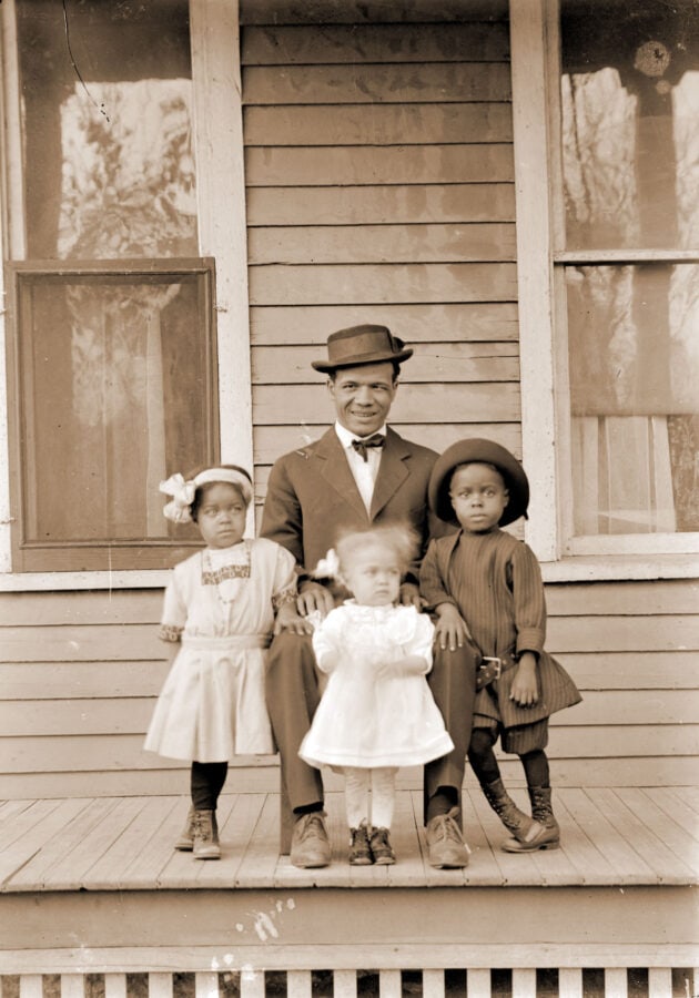 An African American man poses with three very young children. The father sits in a chair, while the three children lean on him.