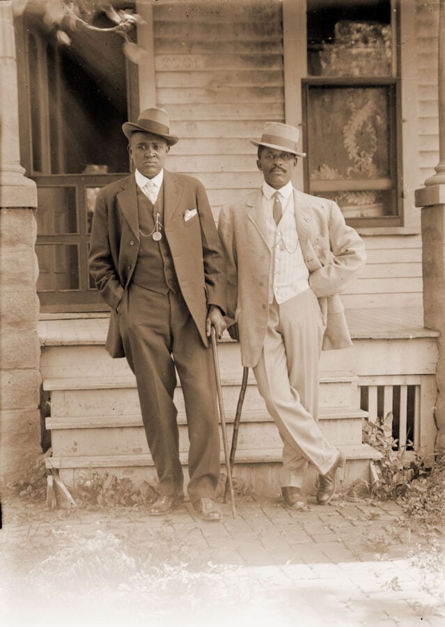 Two African American men stand in front of a house. They are both well-dressed and lean on canes, though the canes appear to be used for fashion instead of medical need.