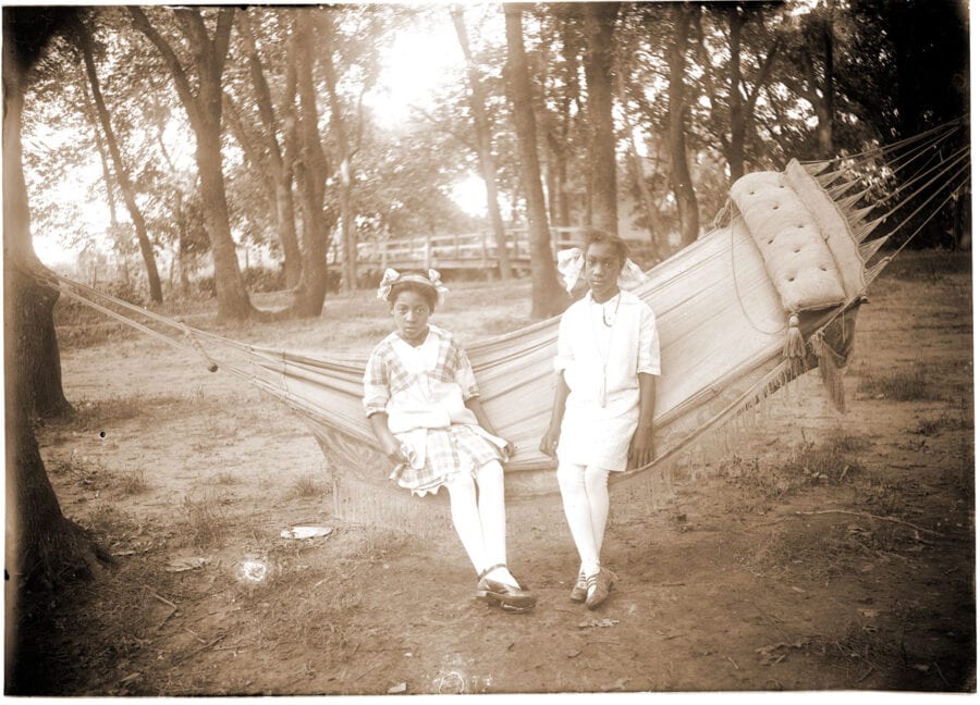 Two young African American girls sit on a hammock outside. There is a bridge and lots of trees behind them.