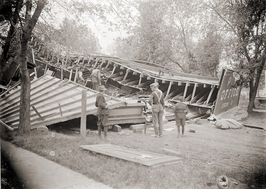 Multiple boys look at a collapsed building. Two men stand in the rubble.