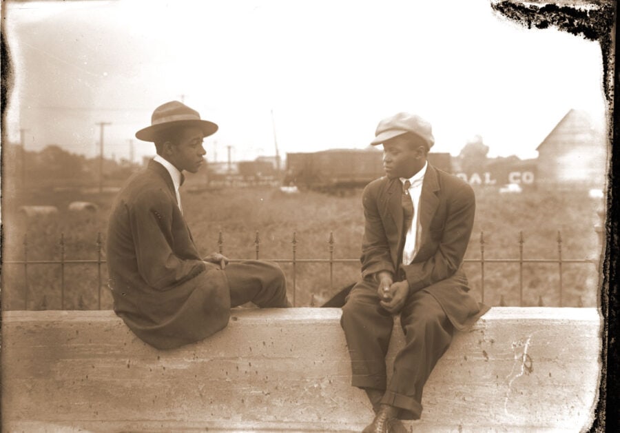 Two teenaged African American men sit on a wall wearing suits with more casual hats. A freight train is visible behind them.
