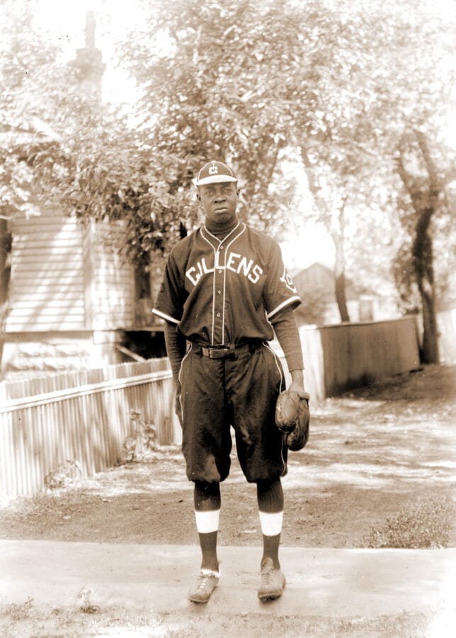 An African American man stands outside in a baseball uniform. The front of the uniform reads "GILLENS."