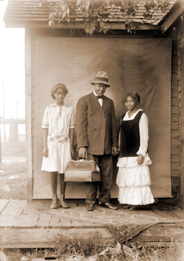 Two young African American women stand outside in front of a backdrop. A rug is at their feet. Between them is a man with a large fedora-like hat carrying a structured bag.