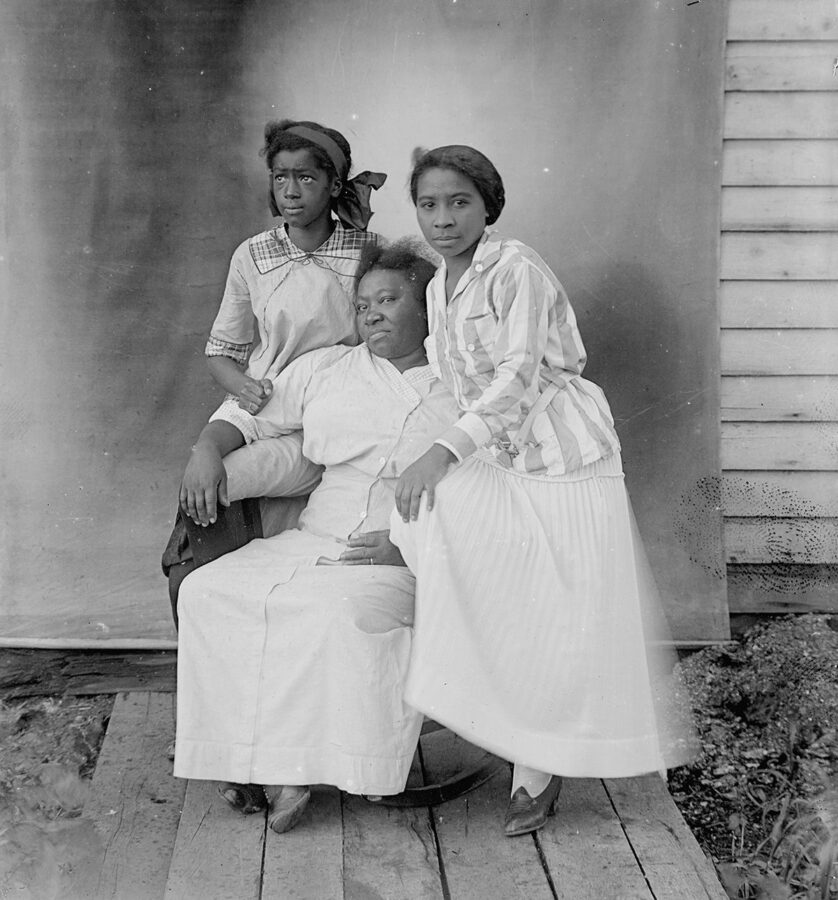 An older African American woman sits in a chair. Two younger African American women stand next to her closely.
