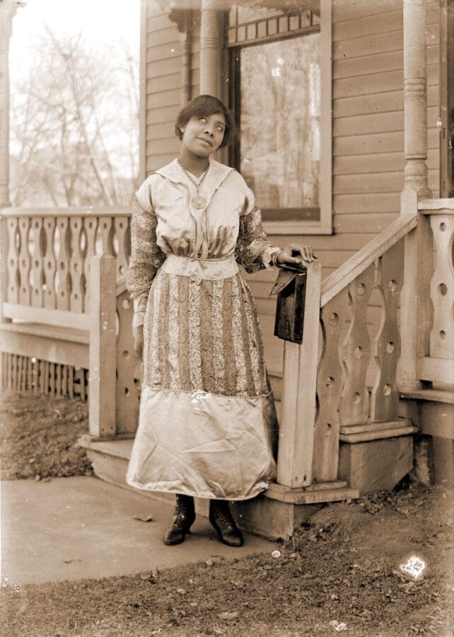 A woman poses at the bottom of her front steps next to a letterbox. She looks up and to the side.