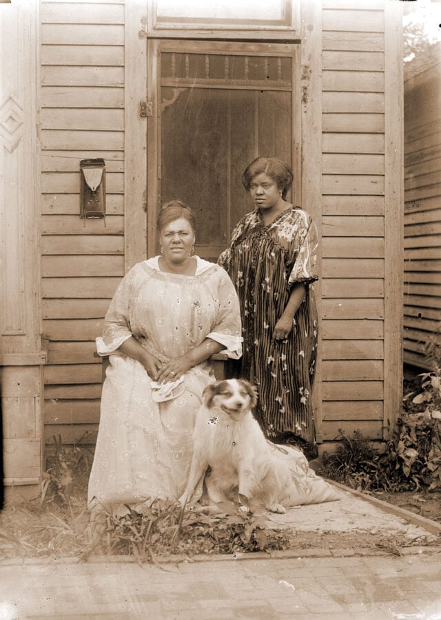 Two African American women pose in front of their house. A dog sits at their feet.
