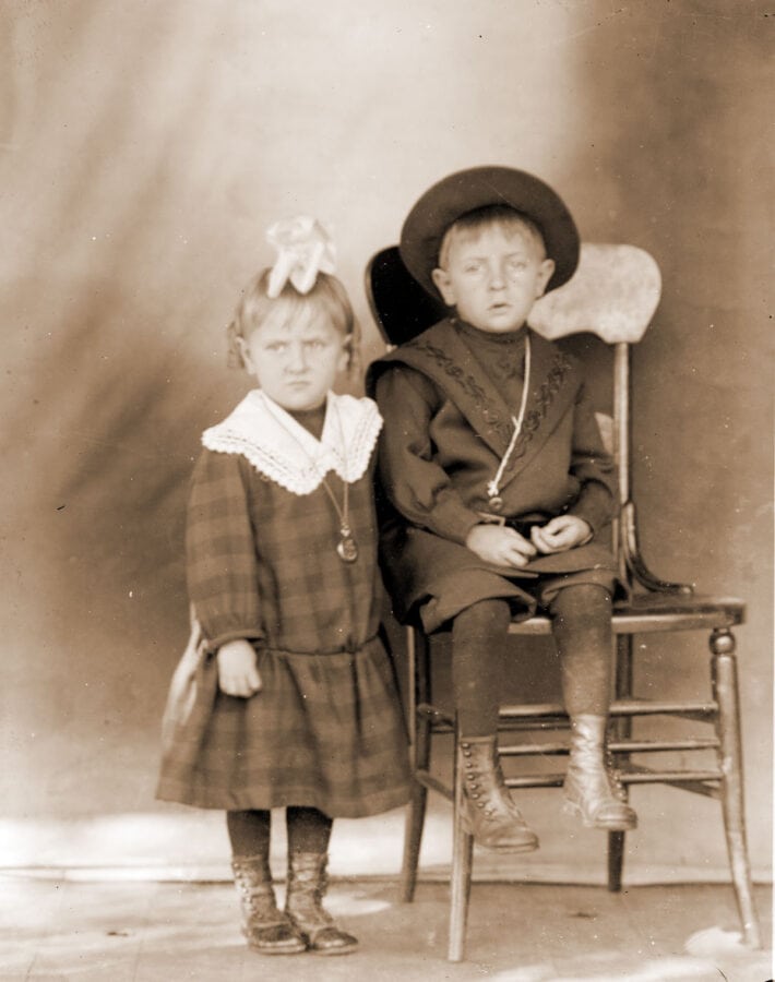Two white, blonde children pose for a picture. The young girl stands next to a chair, upon which a young boy sits.